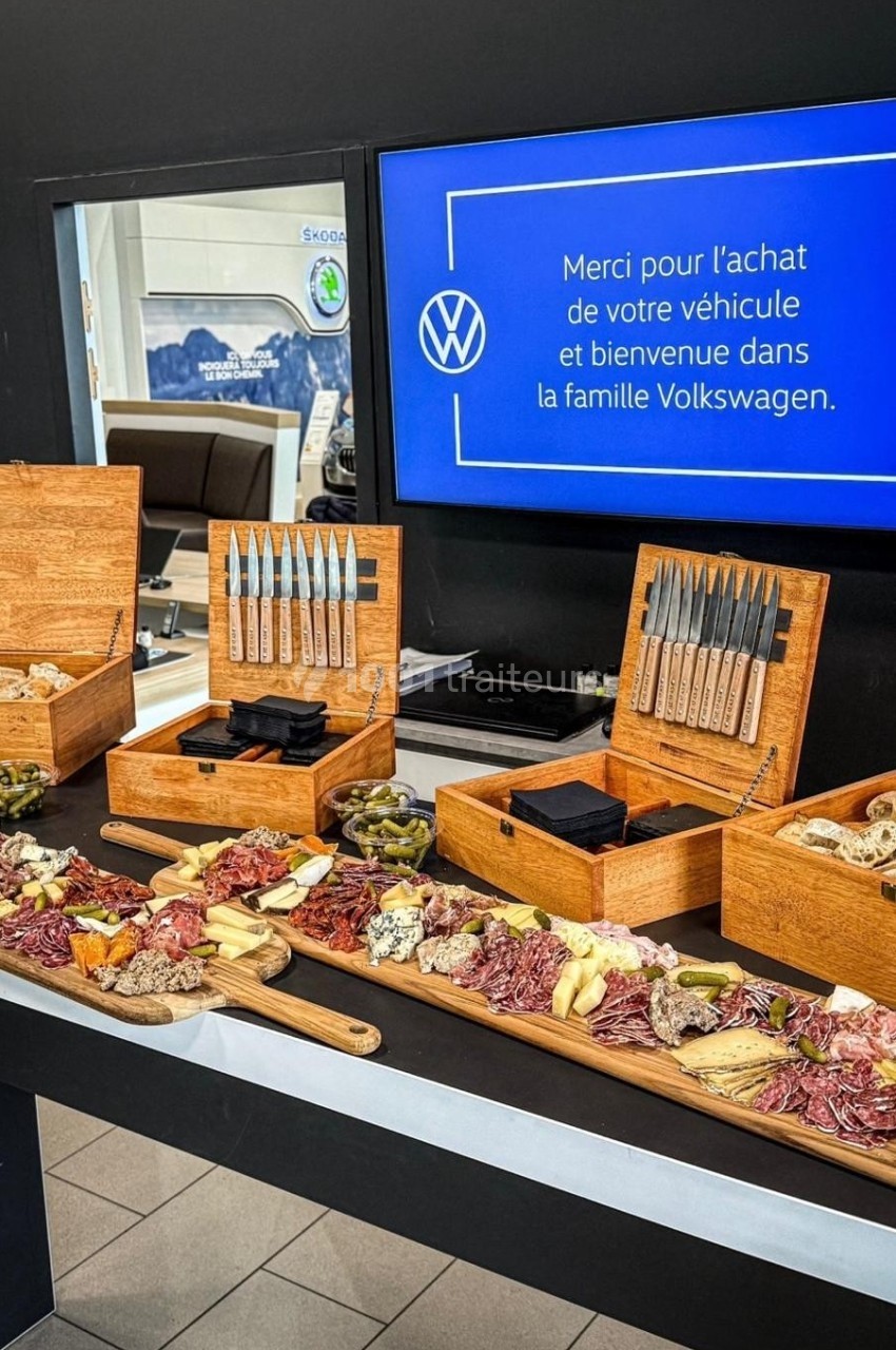 Plateau de charcuterie et fromages disposé sur une table, avec des couteaux dans des boîtes en bois, devant un écran…
