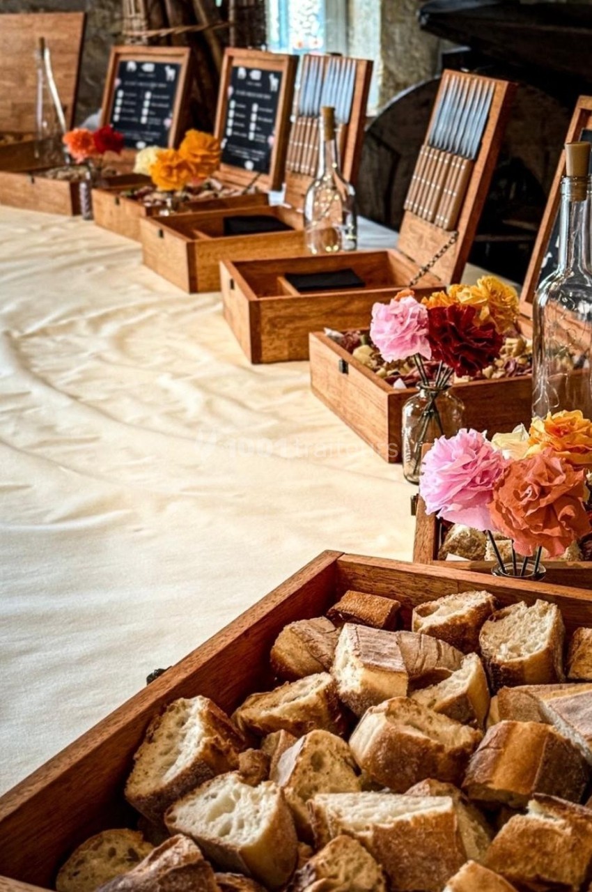 Plateau de pain coupé en morceaux au premier plan, avec une table décorée de fleurs et boîtes en bois en arrière-plan.