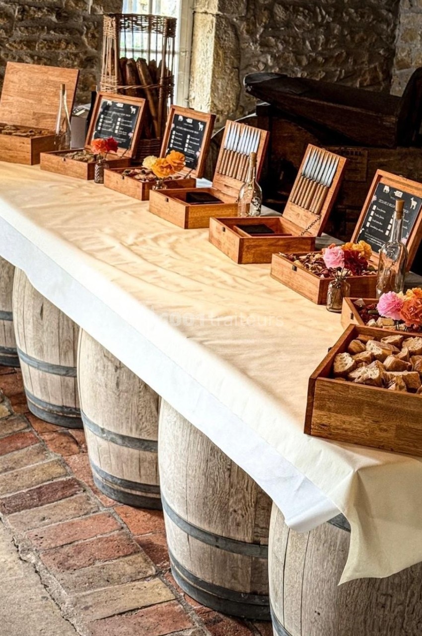 Table dressée sur des tonneaux avec nappes blanches, contenant des boîtes en bois garnies de pains et condiments.