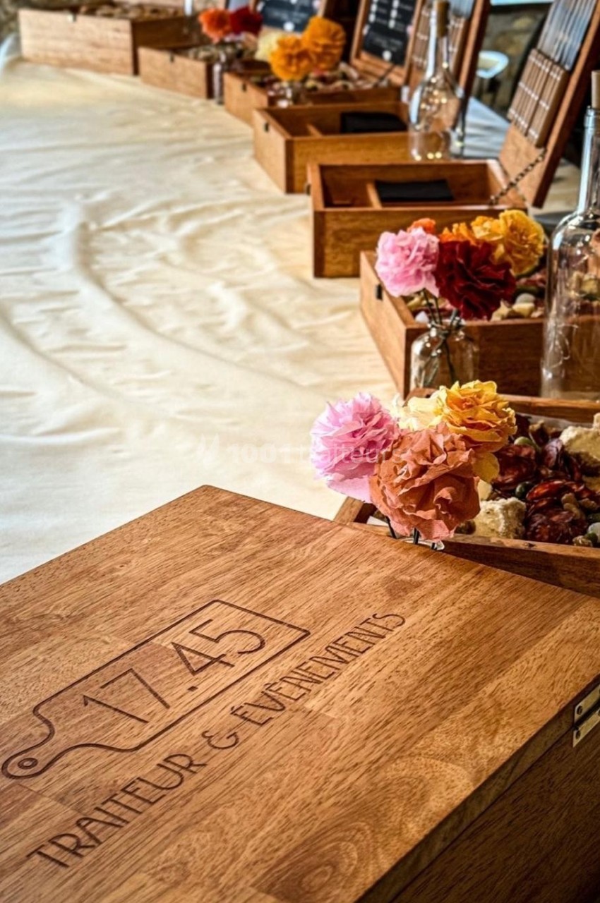 Boîtes en bois gravées ’1745 Traiteur & Événements’ alignées sur une table, décorées de fleurs colorées.