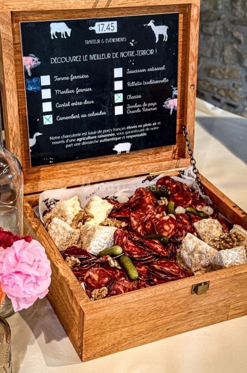 Plateau en bois ouvert contenant des charcuteries, fromages et cornichons, posé sur une table avec une fleur en décoration.