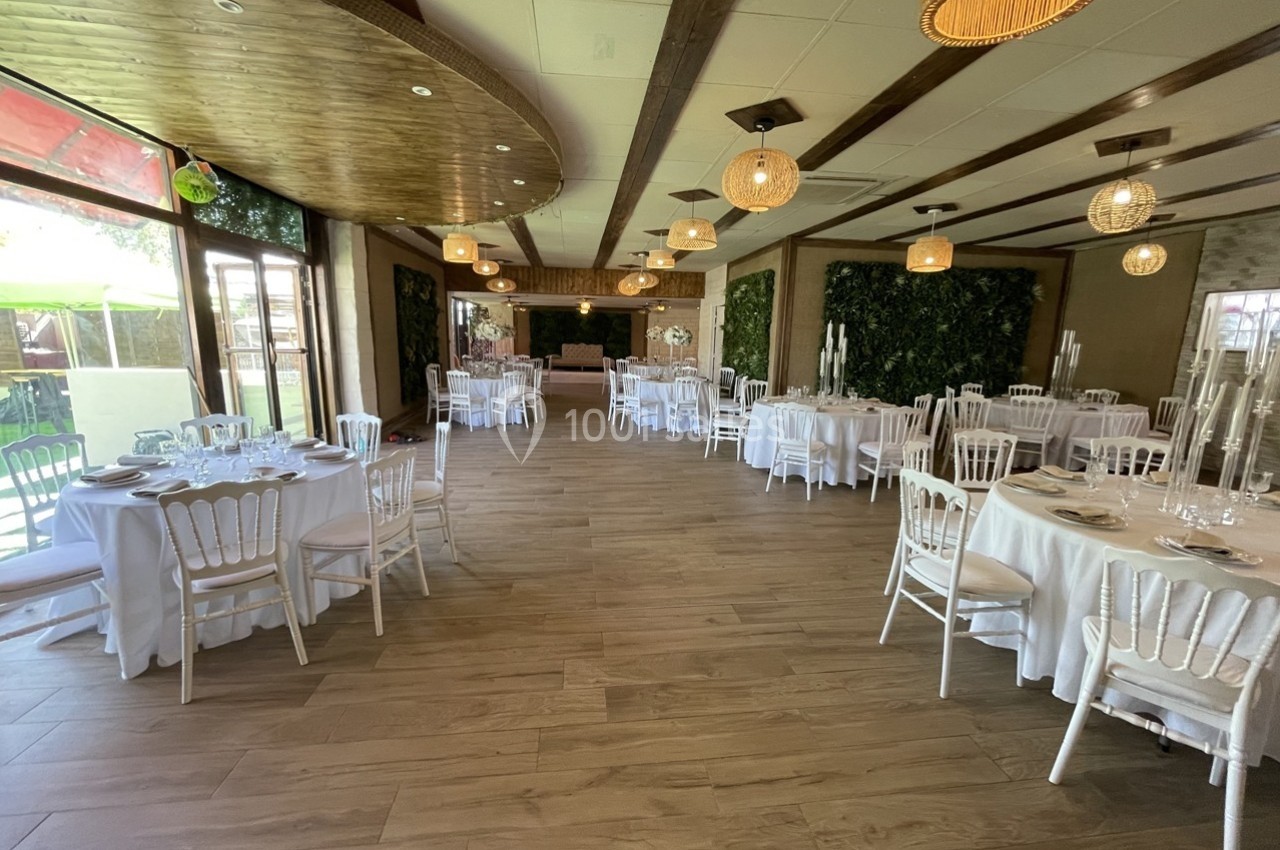 Salle de réception lumineuse avec tables rondes dressées, chaises blanches et décorations végétales sur les murs.