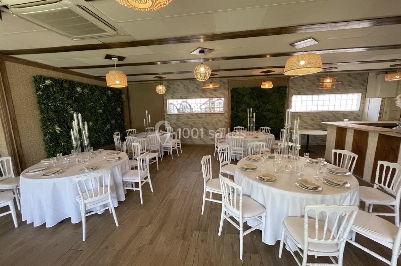 Salle de réception lumineuse avec tables rondes dressées, chaises blanches et murs décorés de végétation.
