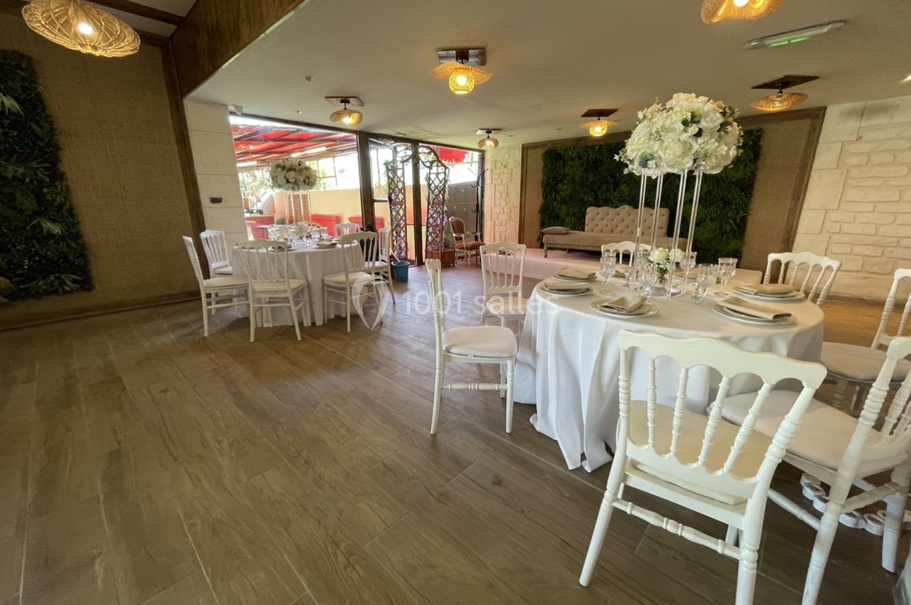 Salle de réception lumineuse avec tables rondes dressées, chaises blanches et décorations florales élégantes.