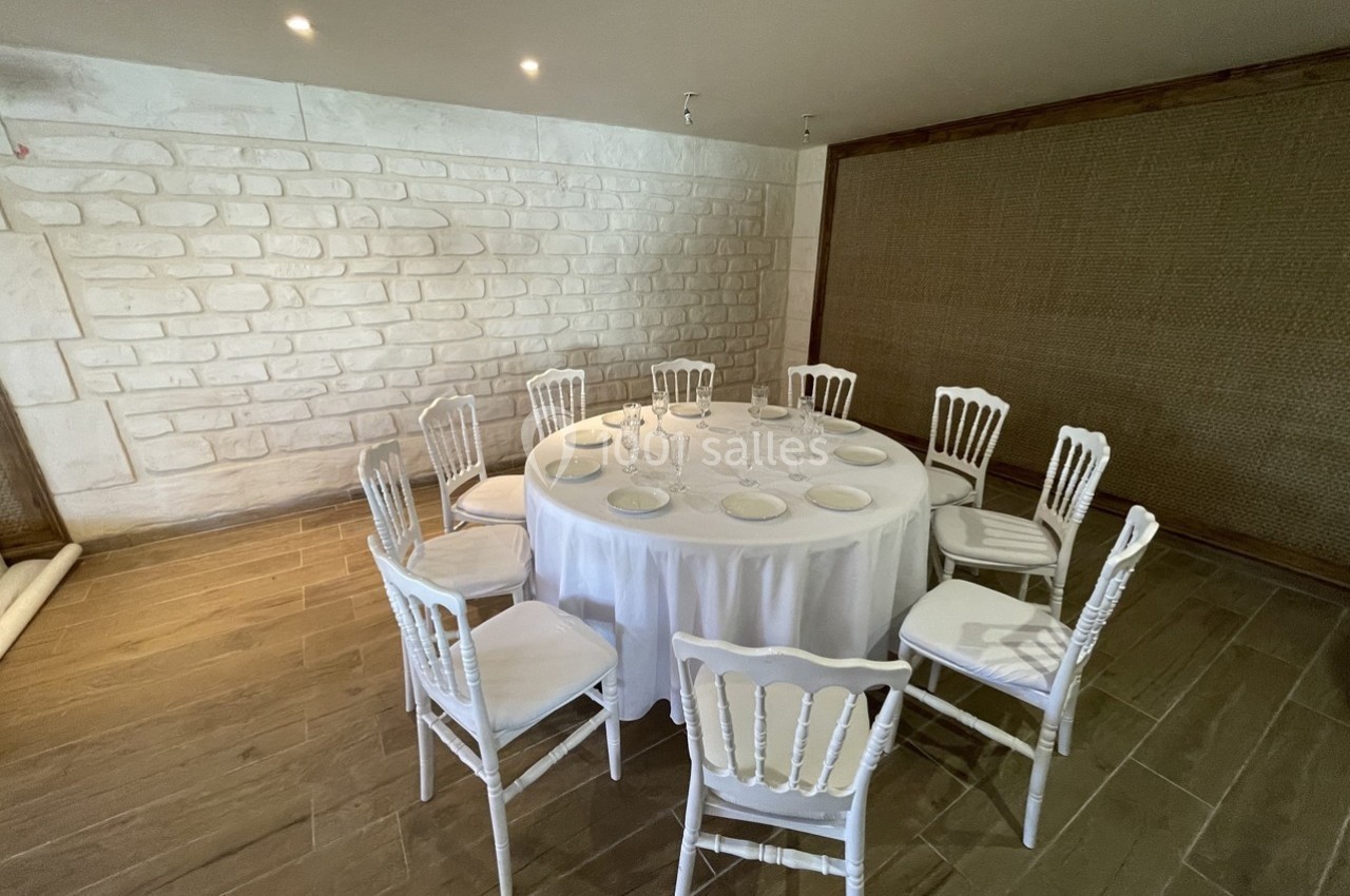 Table ronde dressée avec une nappe blanche, entourée de chaises blanches dans une salle au mur en pierre et parquet clair.