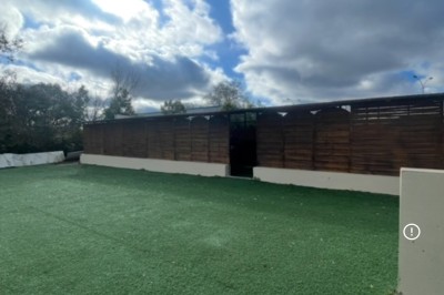 Entrée d'un bâtiment beige avec une toiture rouge, entourée de palissades en bois et d'une pelouse artificielle.