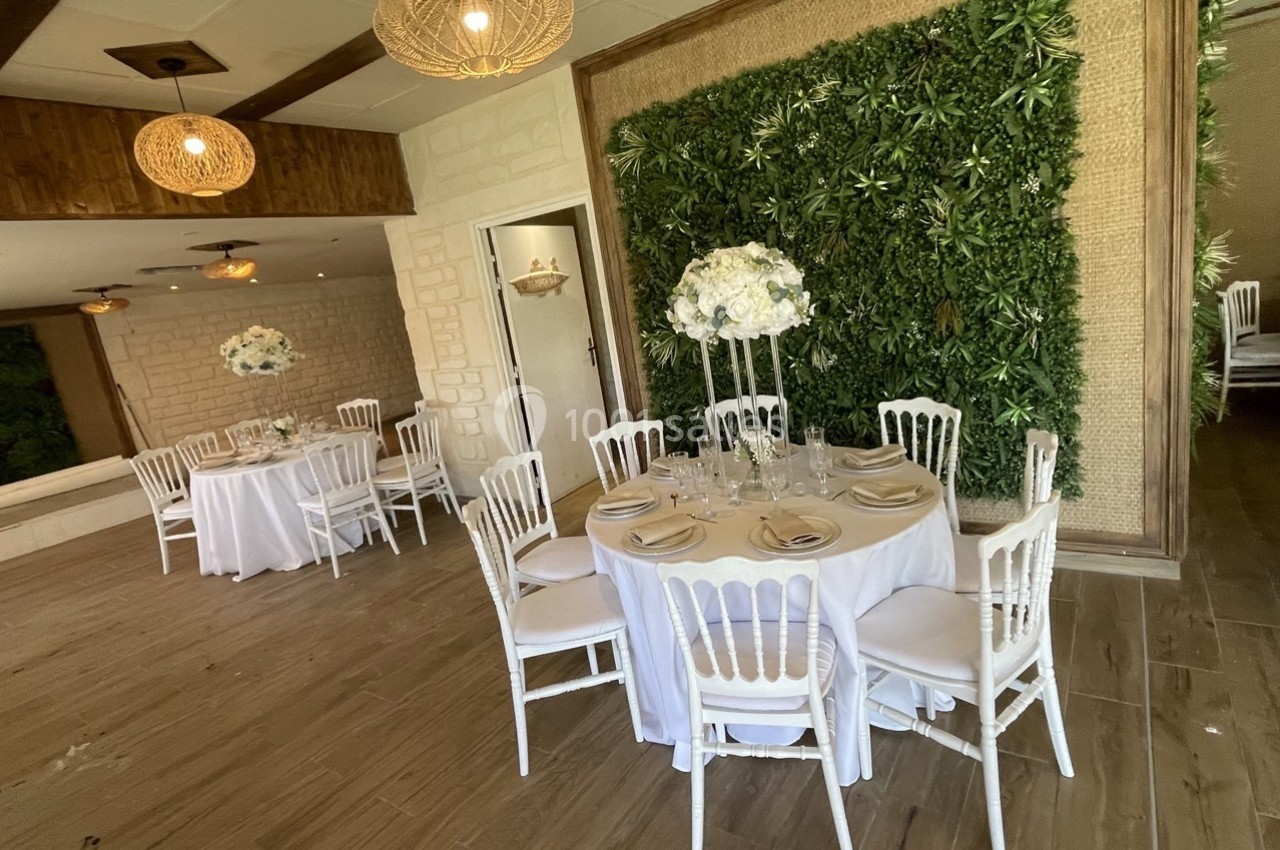 Salle de réception décorée avec des tables rondes, nappes blanches, chaises élégantes et mur végétal en arrière-plan.