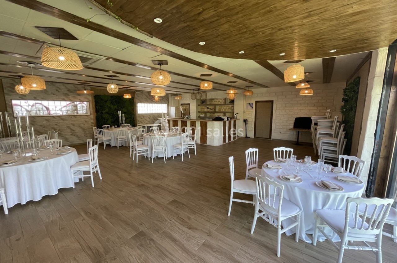 Salle de réception lumineuse avec tables rondes dressées, chaises blanches et décorations en bois et osier.