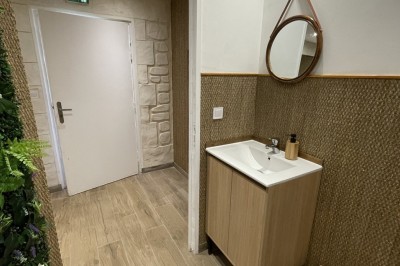 Entrée d'un bâtiment beige avec une toiture rouge, entourée de palissades en bois et d'une pelouse artificielle.