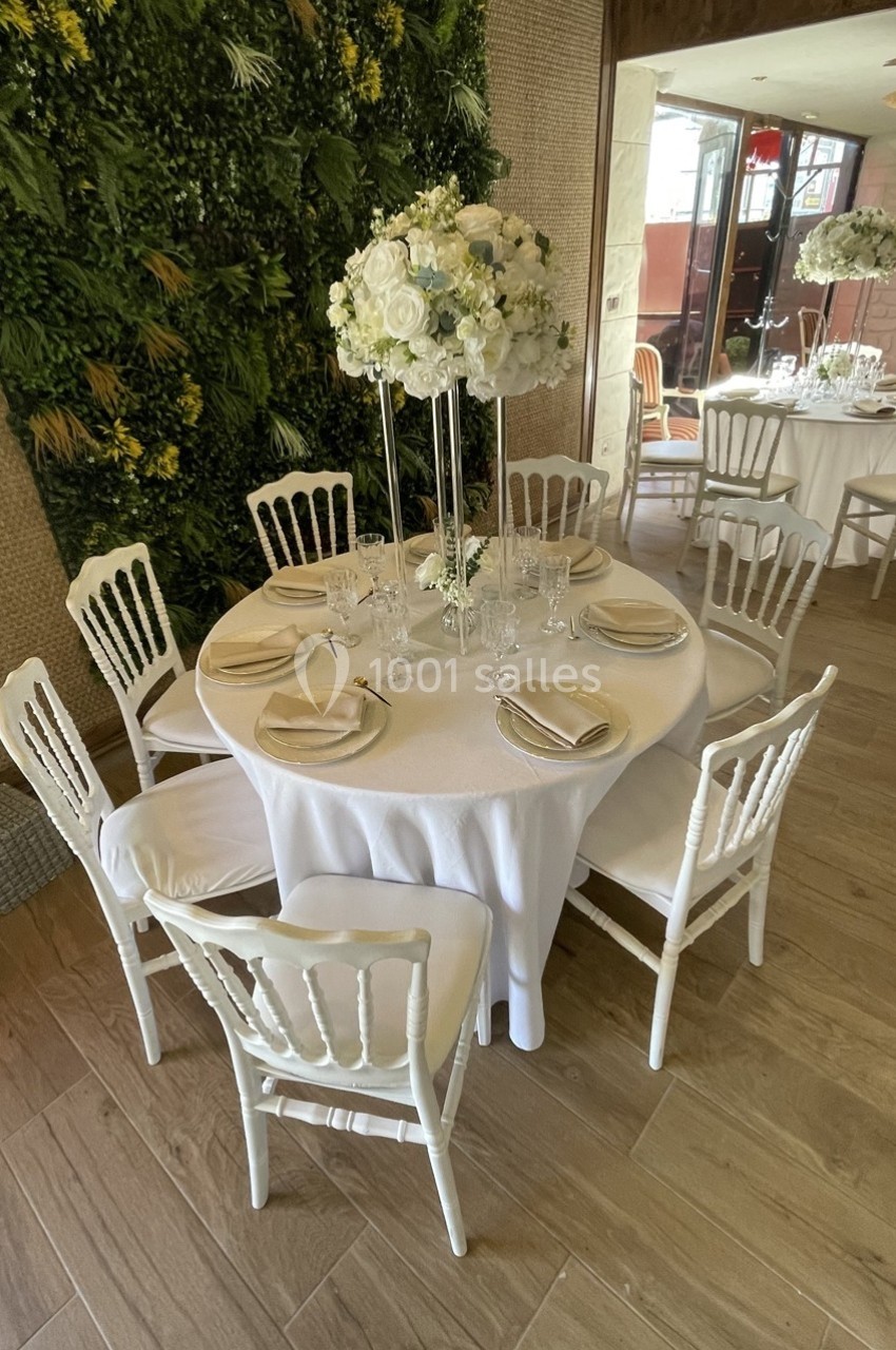 Table ronde décorée pour un événement, avec nappes blanches, chaises assorties et centre de table floral élégant.