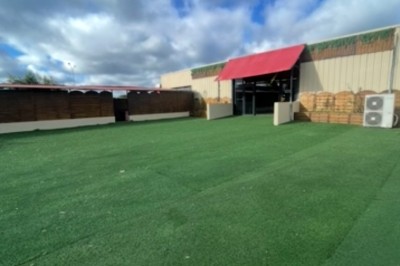 Entrée d'un bâtiment beige avec une toiture rouge, entourée de palissades en bois et d'une pelouse artificielle.