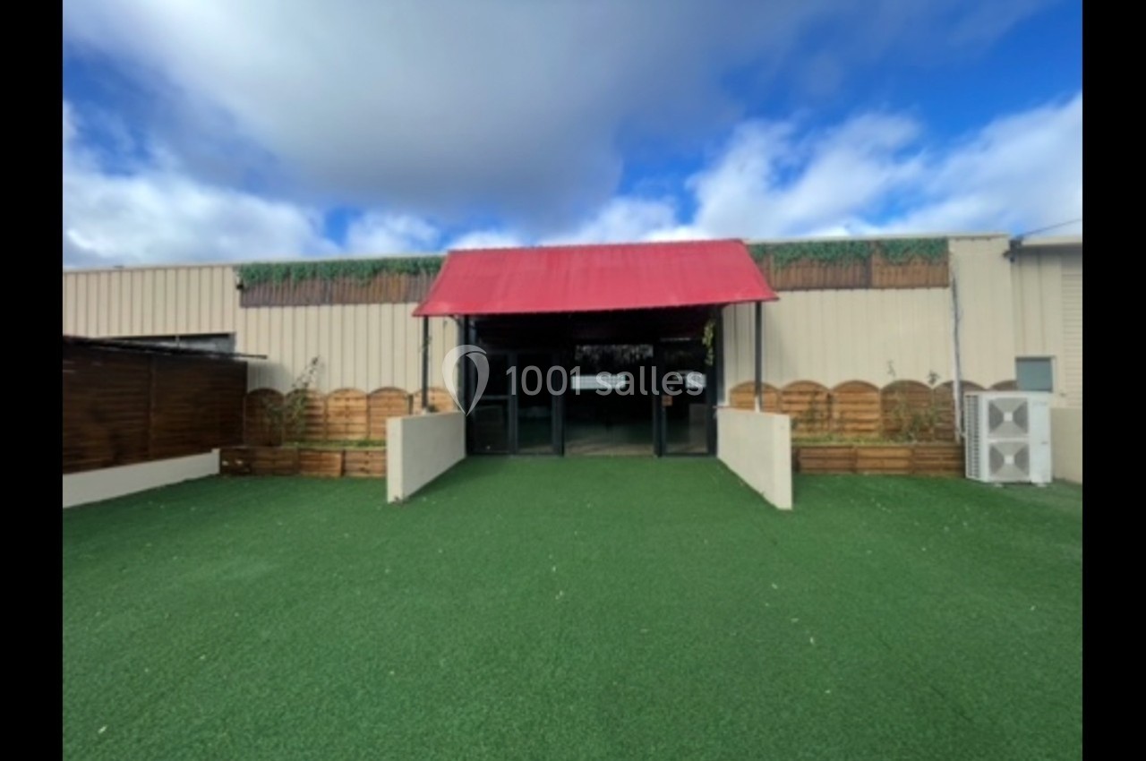 Entrée d'un bâtiment beige avec une toiture rouge, entourée de palissades en bois et d'une pelouse artificielle.