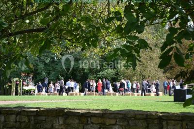 Vue depuis une porte vitrée sur un jardin avec des tables hautes et des chaises disposées près d'un bâtiment en pierre.