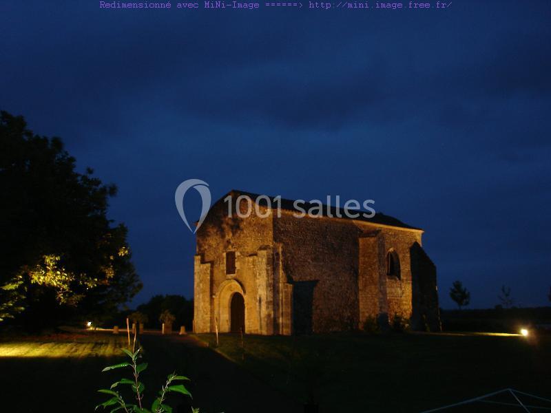 Église en pierre éclairée de nuit, entourée de végétation et d'un ciel sombre.