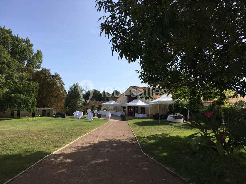 Allée bordée de verdure menant à un bâtiment en pierre avec des tentes blanches installées sur une pelouse.
