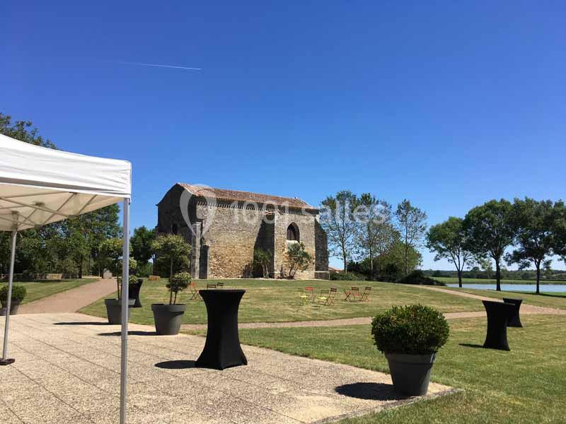 Vue d'une petite chapelle en pierre entourée de verdure, avec des tables hautes et un chapiteau au premier plan.