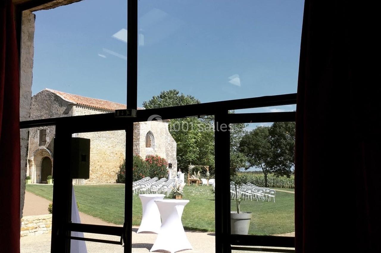 Vue depuis une porte vitrée sur un jardin avec des tables hautes et des chaises disposées près d'un bâtiment en pierre.