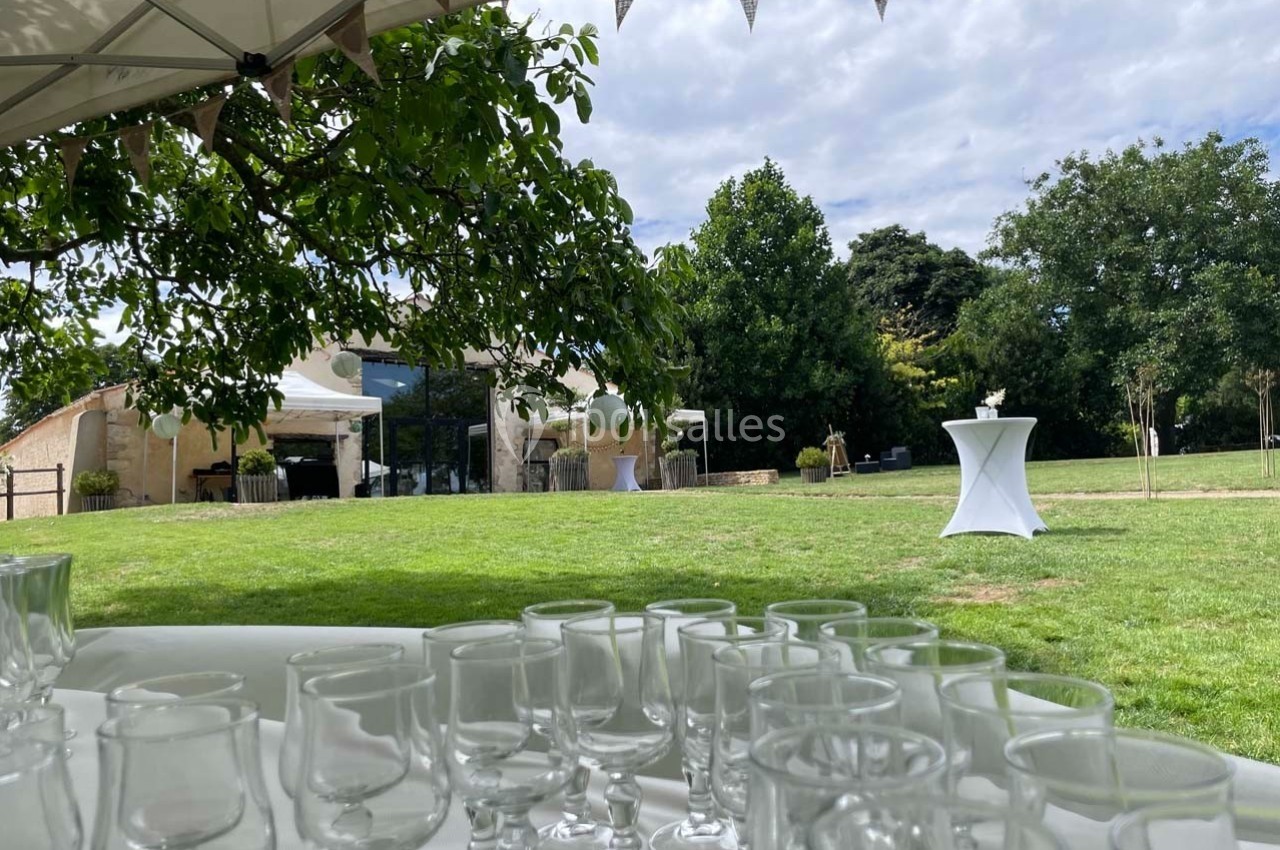 Verres disposés sur une table sous un chapiteau, avec vue sur un jardin verdoyant et un bâtiment en arrière-plan.