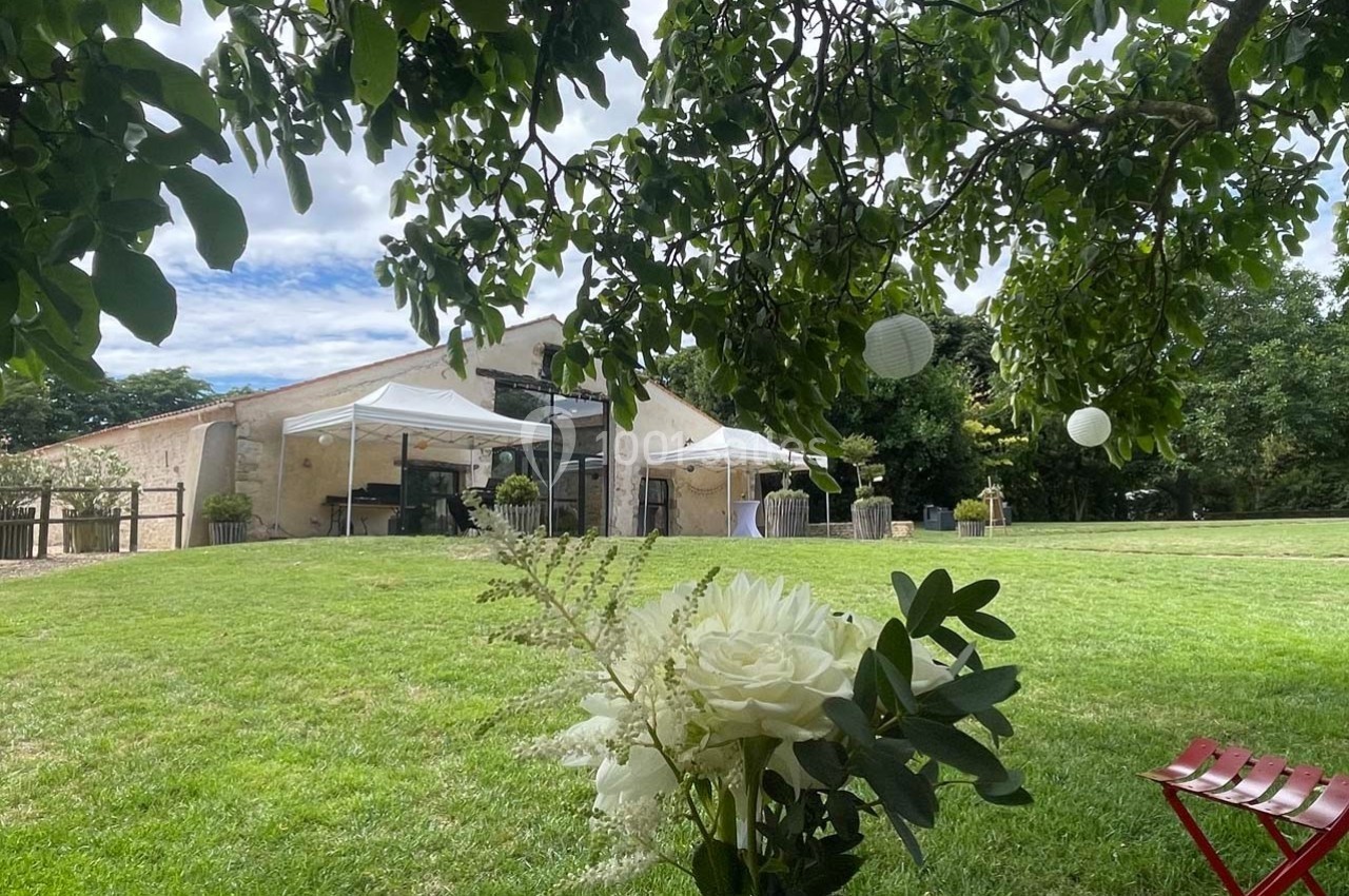 Vue d'un jardin avec pelouse, barnum blanc, bâtiment en pierre et fleurs au premier plan sous un arbre.