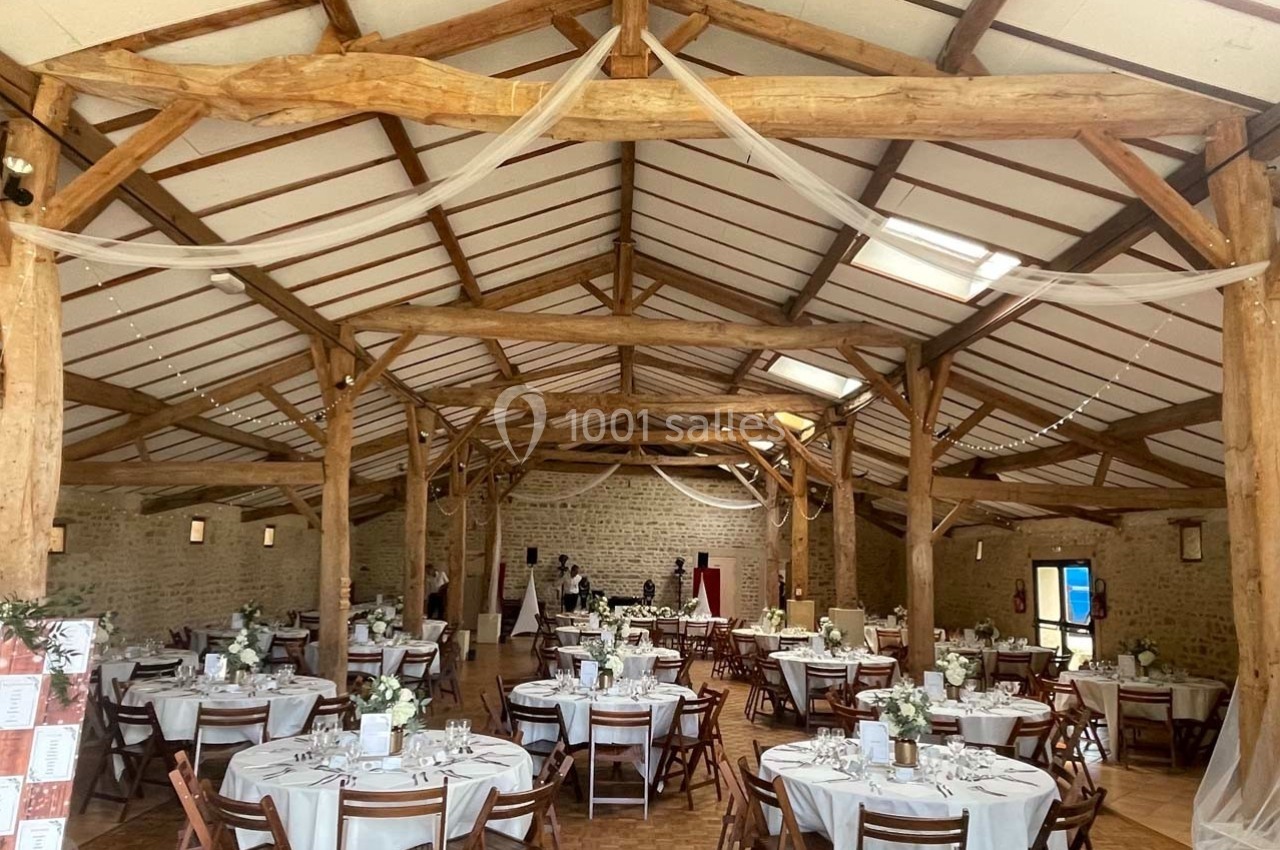 Salle de réception en bois avec tables rondes dressées, décorations blanches et lumière naturelle.