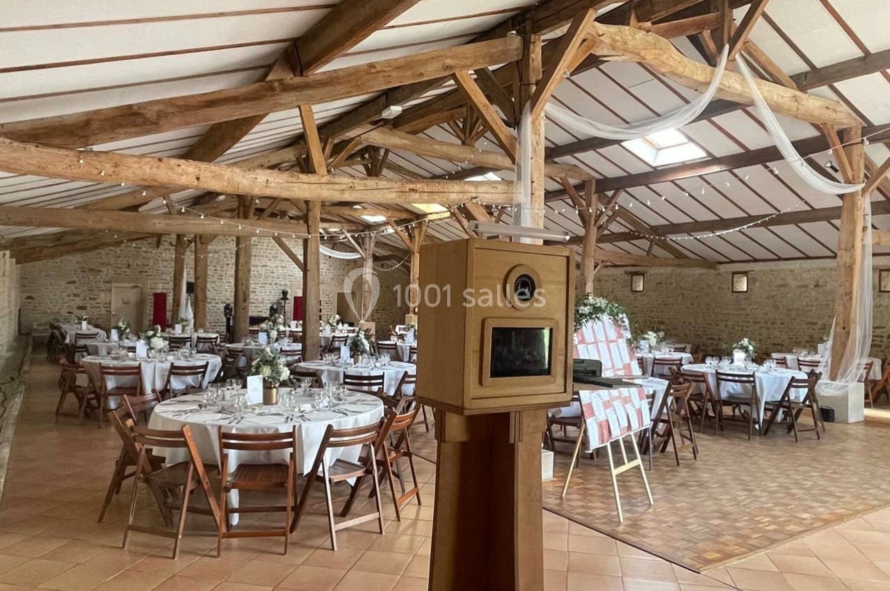 Salle de réception avec tables rondes décorées pour un événement, poutres apparentes et borne photo au premier plan.