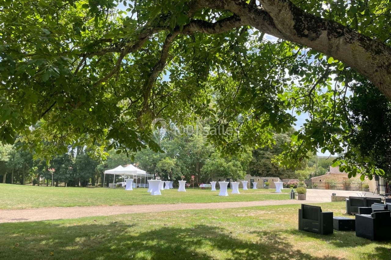 Jardin verdoyant avec tables hautes blanches sous des arbres, une tente et des fauteuils noirs en arrière-plan.