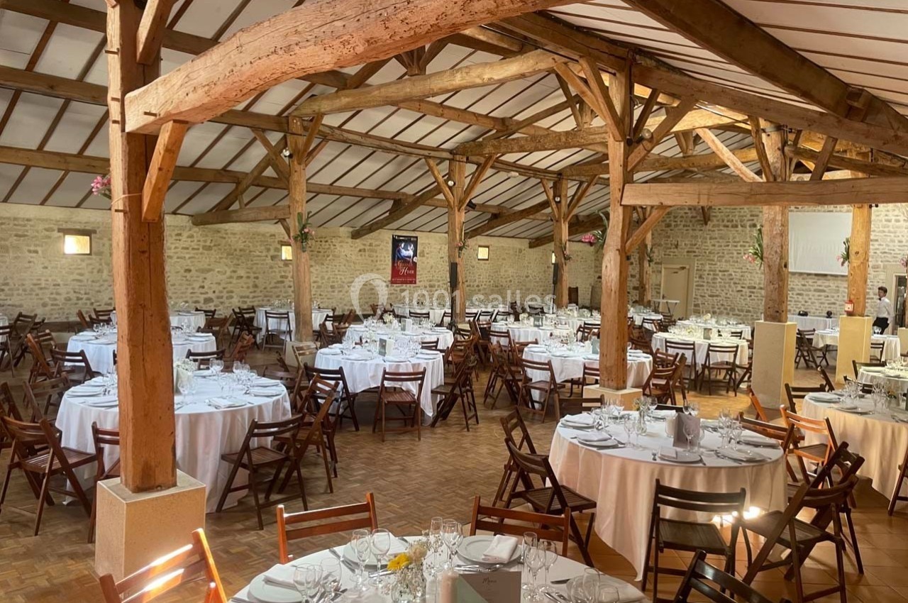 Salle de réception avec poutres apparentes, tables rondes dressées avec nappes blanches et chaises en bois.