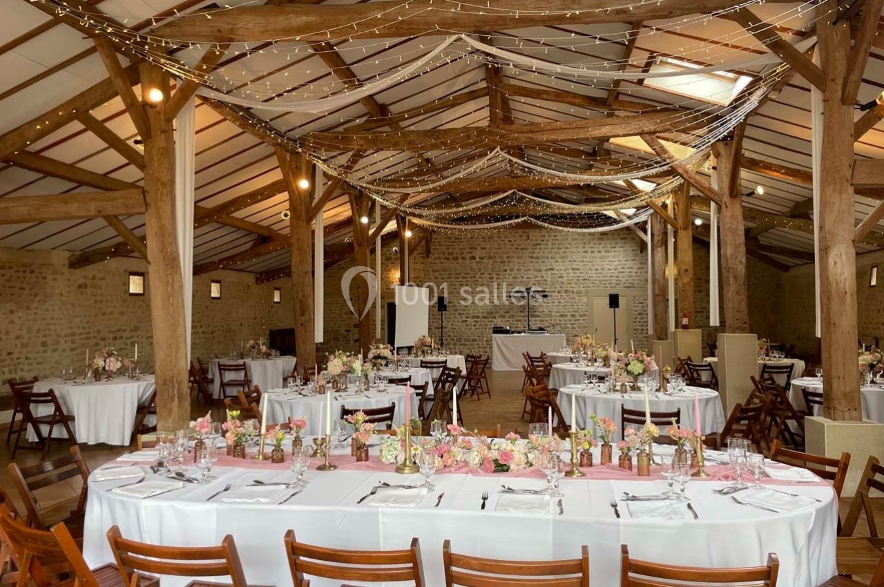 Salle de réception rustique avec poutres apparentes, tables décorées de fleurs et guirlandes lumineuses suspendues.