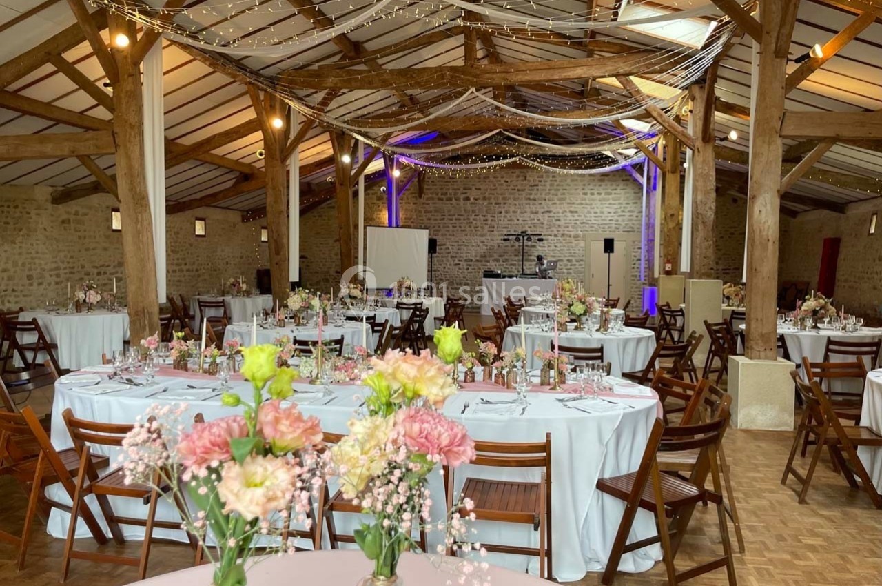 Salle de réception rustique avec tables décorées de fleurs pastel, chaises en bois et guirlandes lumineuses suspendues.