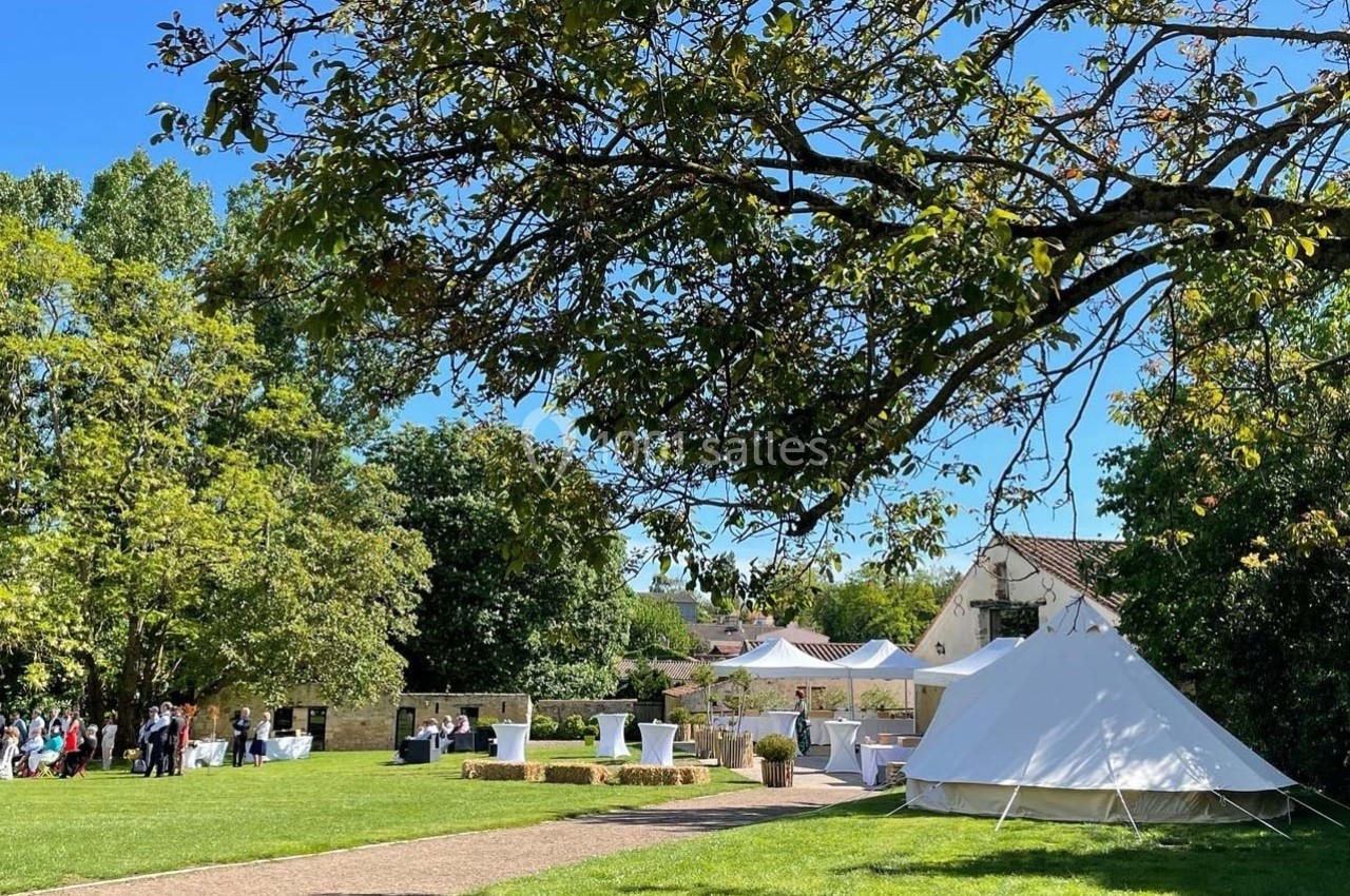 Jardin verdoyant avec une tente blanche, des tables et des invités rassemblés sous un ciel bleu ensoleillé.