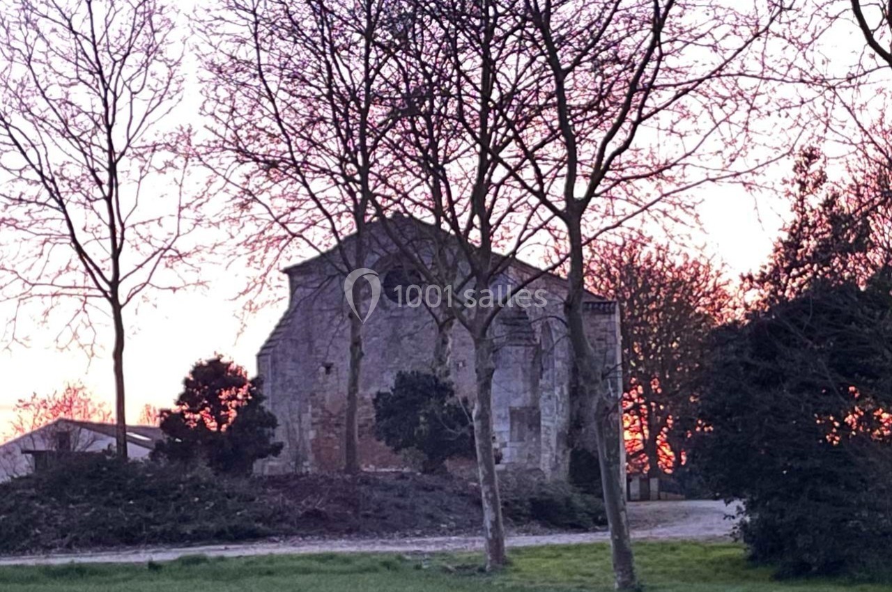 Vieille bâtisse en pierre entourée d'arbres dénudés, avec un ciel de coucher de soleil en arrière-plan.