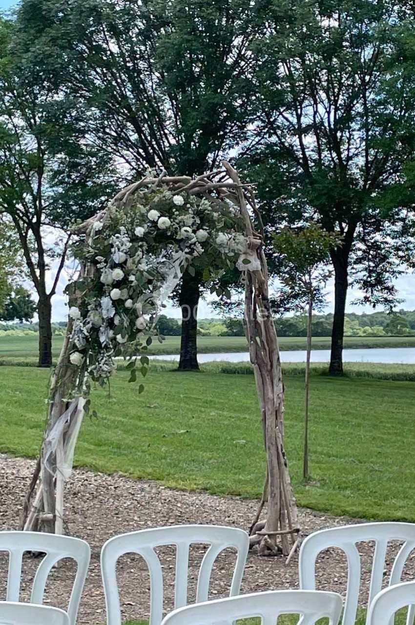 Arche en bois décorée de fleurs blanches dans un cadre extérieur verdoyant, avec des chaises blanches disposées devant.
