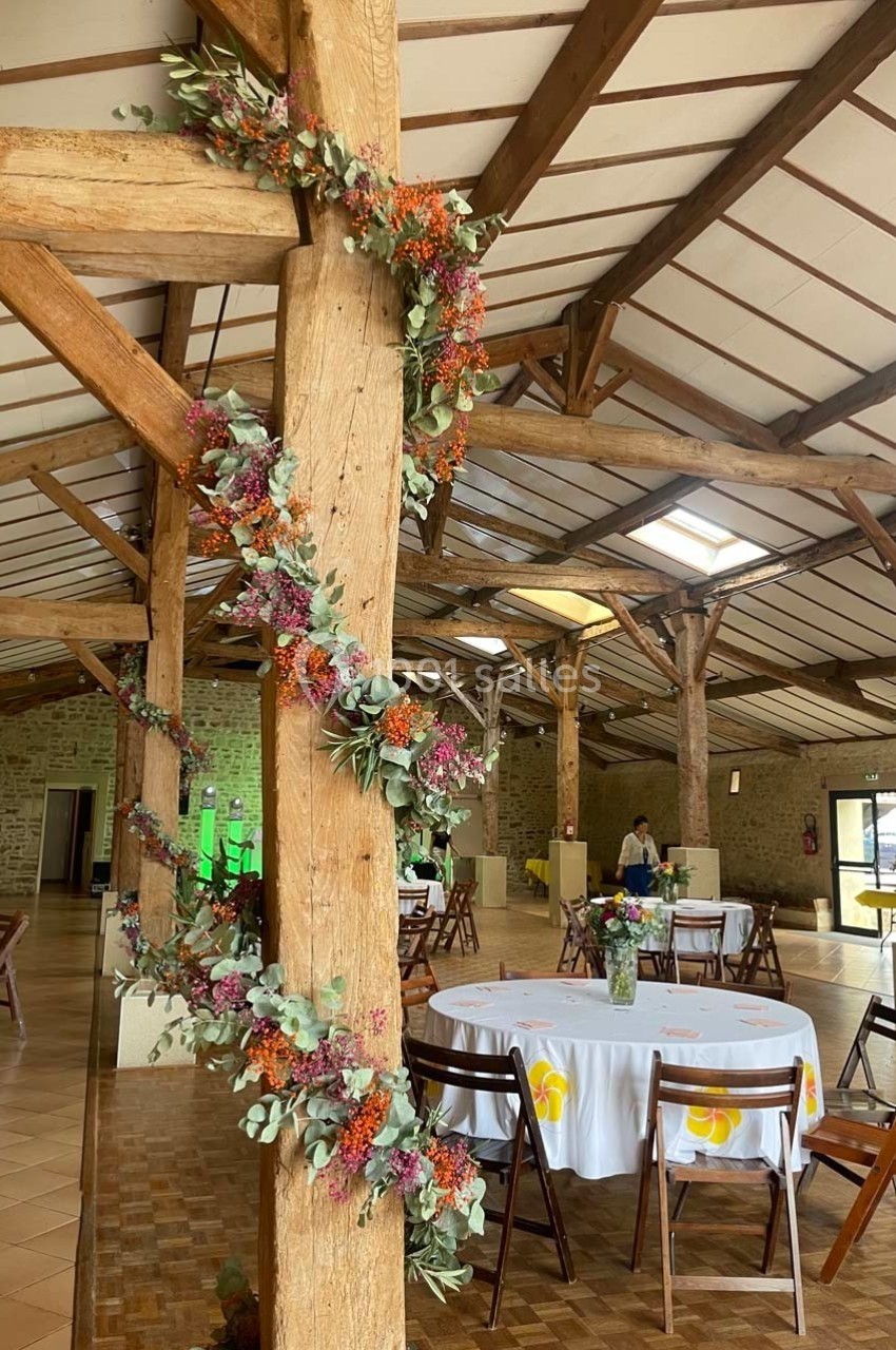 Salle de réception avec poutres en bois décorées de guirlandes florales, tables rondes dressées et chaises en bois.
