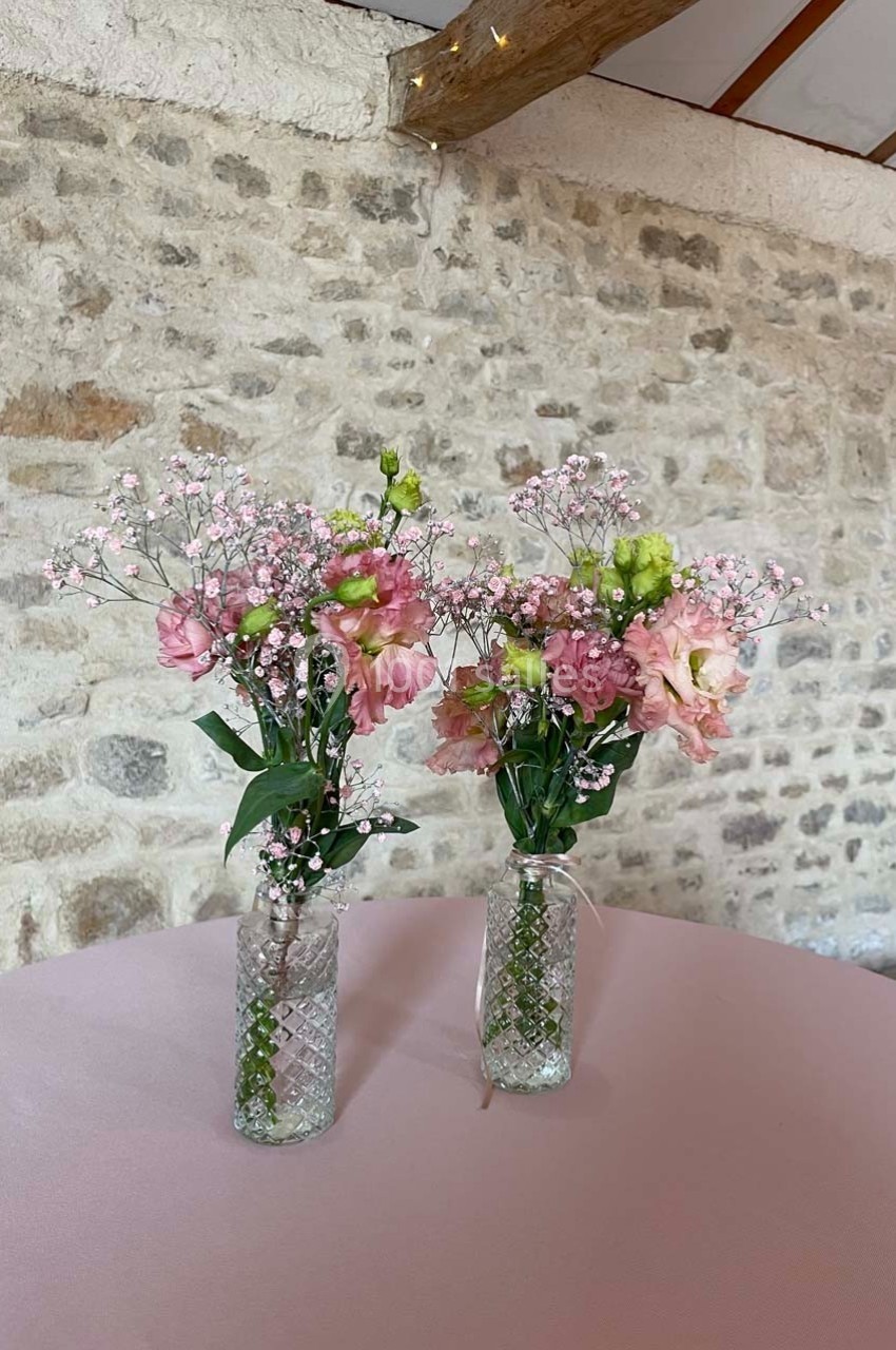 Deux vases en verre contenant des bouquets de fleurs roses et blanches, posés sur une table rose devant un mur en pierre.
