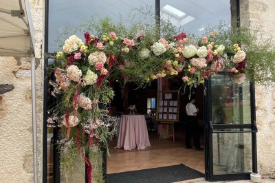 Miniature Location salle Le Bernard (Vendée) - La Grange du Prieuré #27 Centre de table avec des fleurs roses et blanches dans un vase doré, entouré de bougies et de verres sur une nappe blanche.