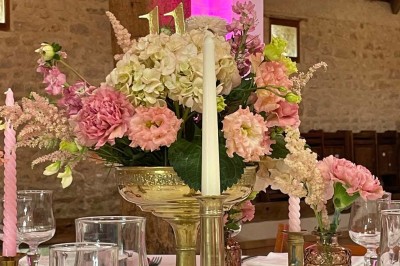 Miniature Location salle Le Bernard (Vendée) - La Grange du Prieuré #28 Centre de table avec des fleurs roses et blanches dans un vase doré, entouré de bougies et de verres sur une nappe blanche.