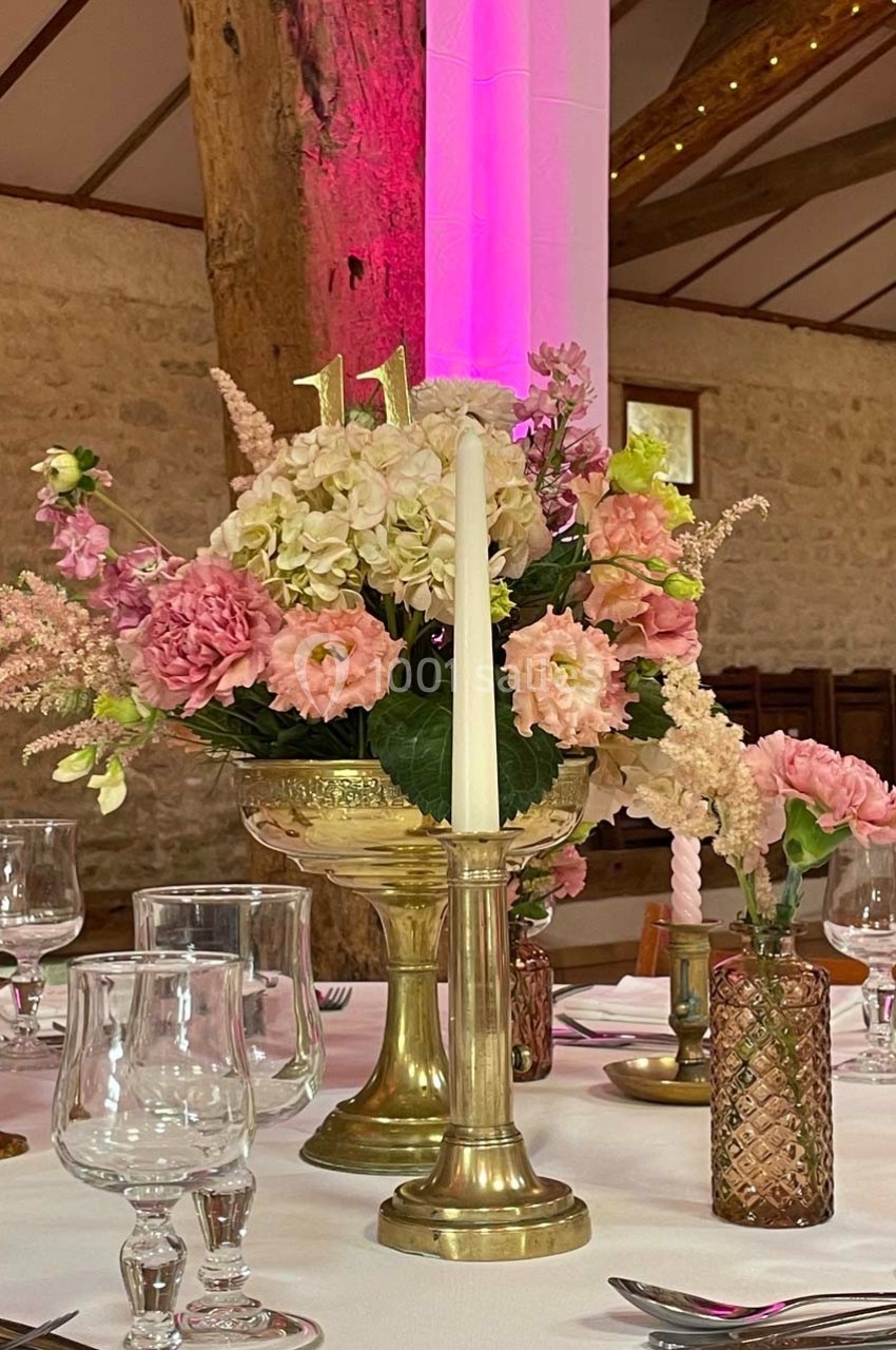 Centre de table avec des fleurs roses et blanches dans un vase doré, entouré de bougies et de verres sur une nappe blanche.