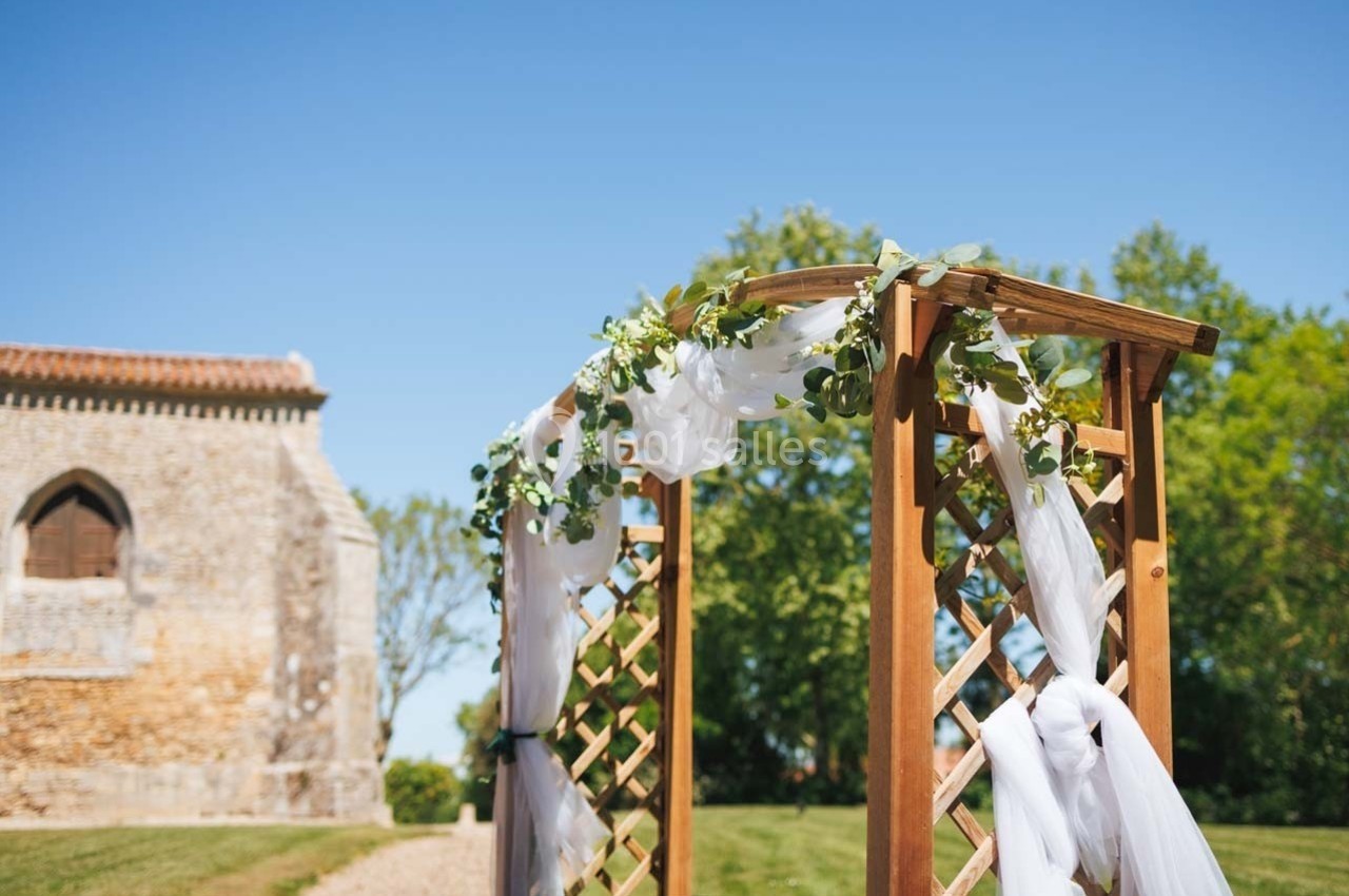 Arche en bois décorée de voilages blancs et de feuillage, installée dans un jardin près d'un bâtiment en pierre.