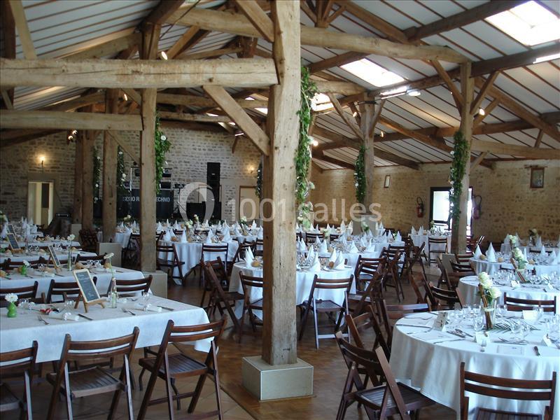 Salle de réception avec poutres apparentes, tables rondes dressées et décorations végétales suspendues.