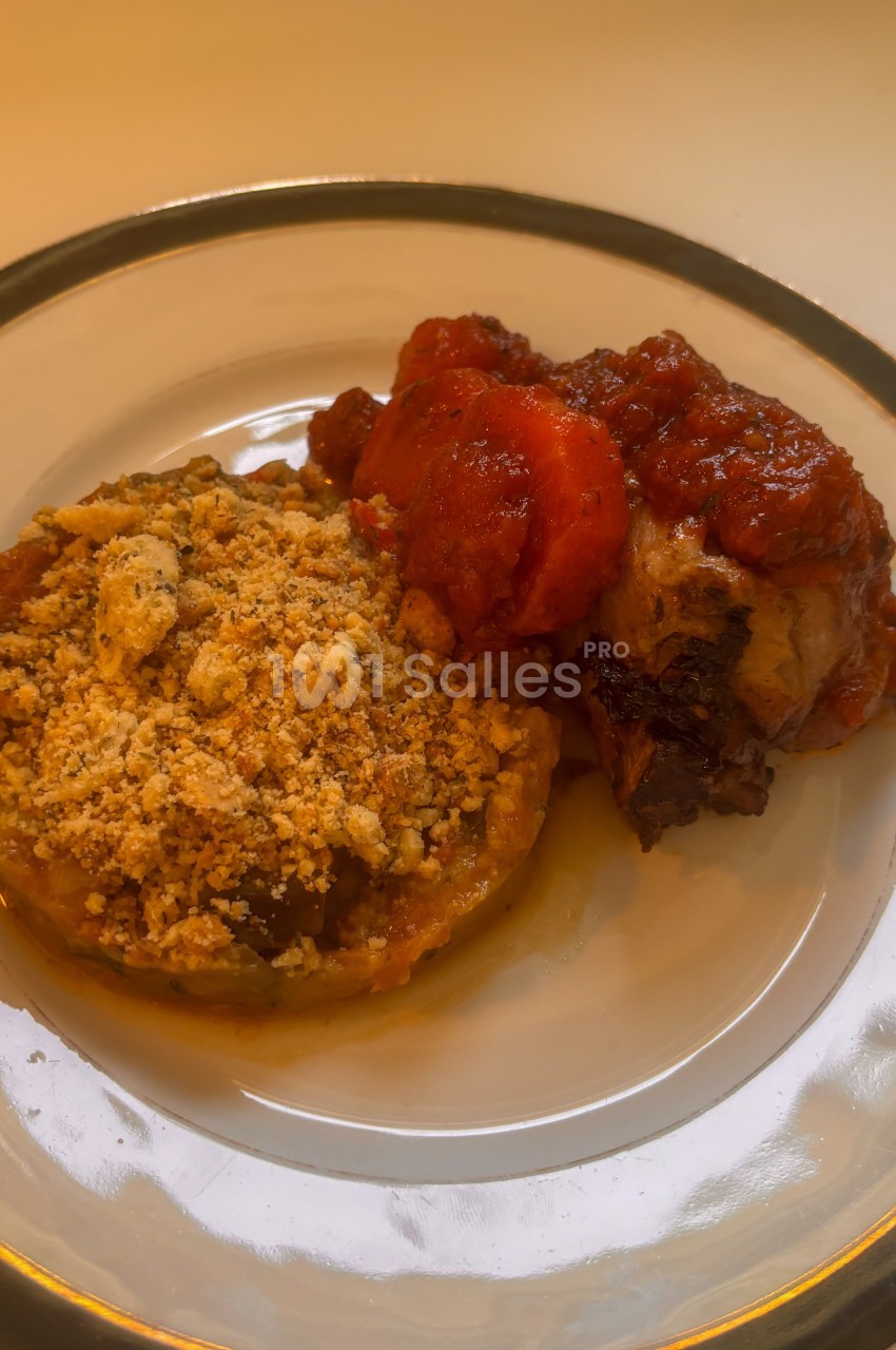 Assiette contenant un gratin de légumes avec une chapelure dorée et un morceau de viande en sauce tomate.