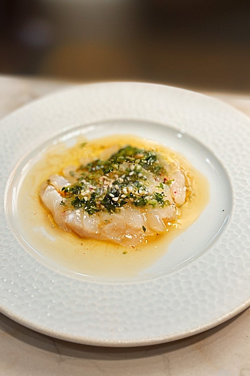 Carpaccio de poisson blanc nappé d'une sauce citronnée, garni de fines herbes et d'épices, servi dans une assiette blanche.