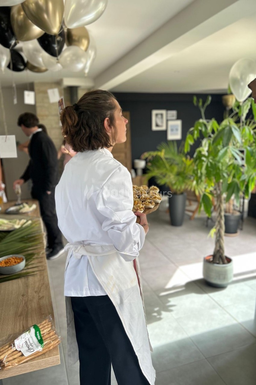 Une personne en blouse blanche tient un plateau de nourriture dans une pièce décorée de ballons et de plantes.
