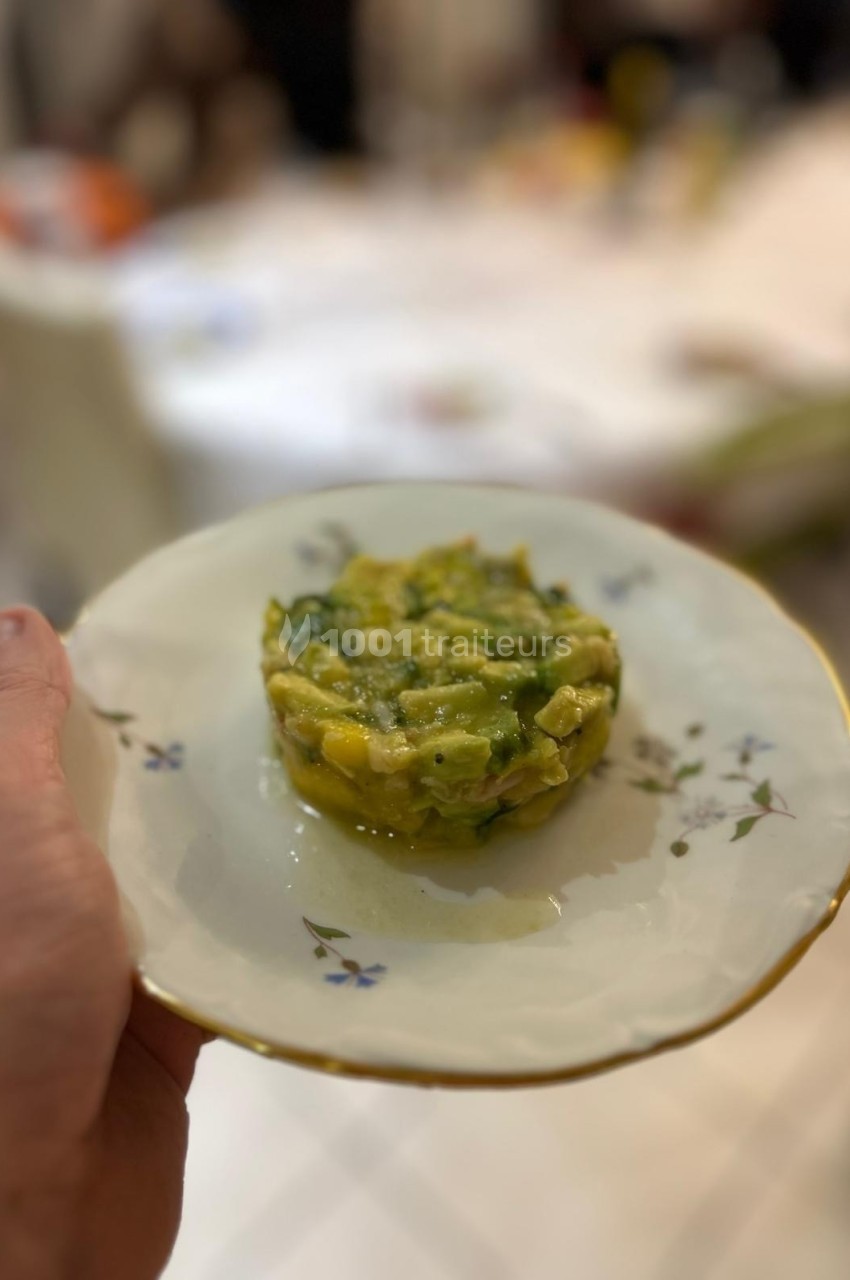 Assiette tenue à la main contenant un tartare d'avocat et de mangue, présentée sur une table en arrière-plan flou.