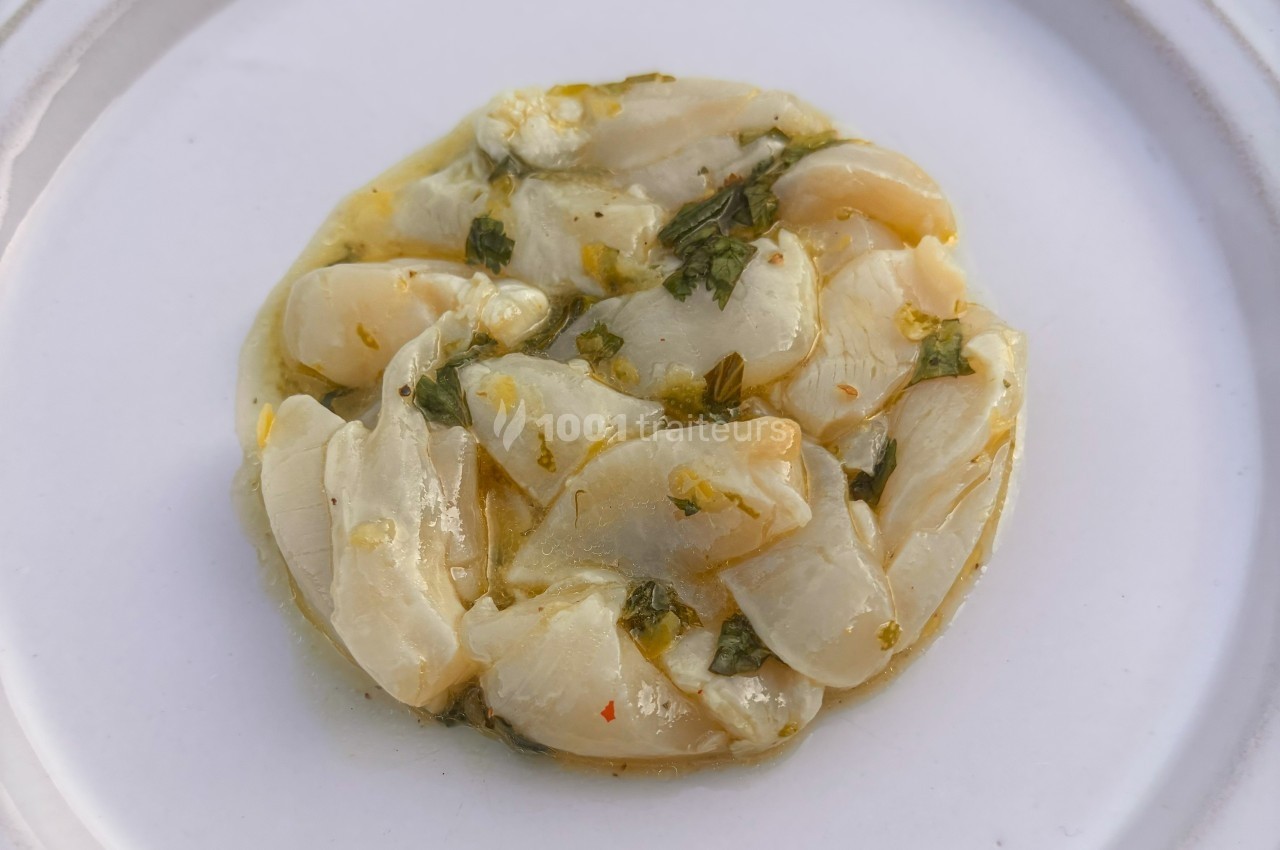 Tranches de noix de Saint-Jacques marinées avec des herbes et de l'huile, présentées sur une assiette blanche.