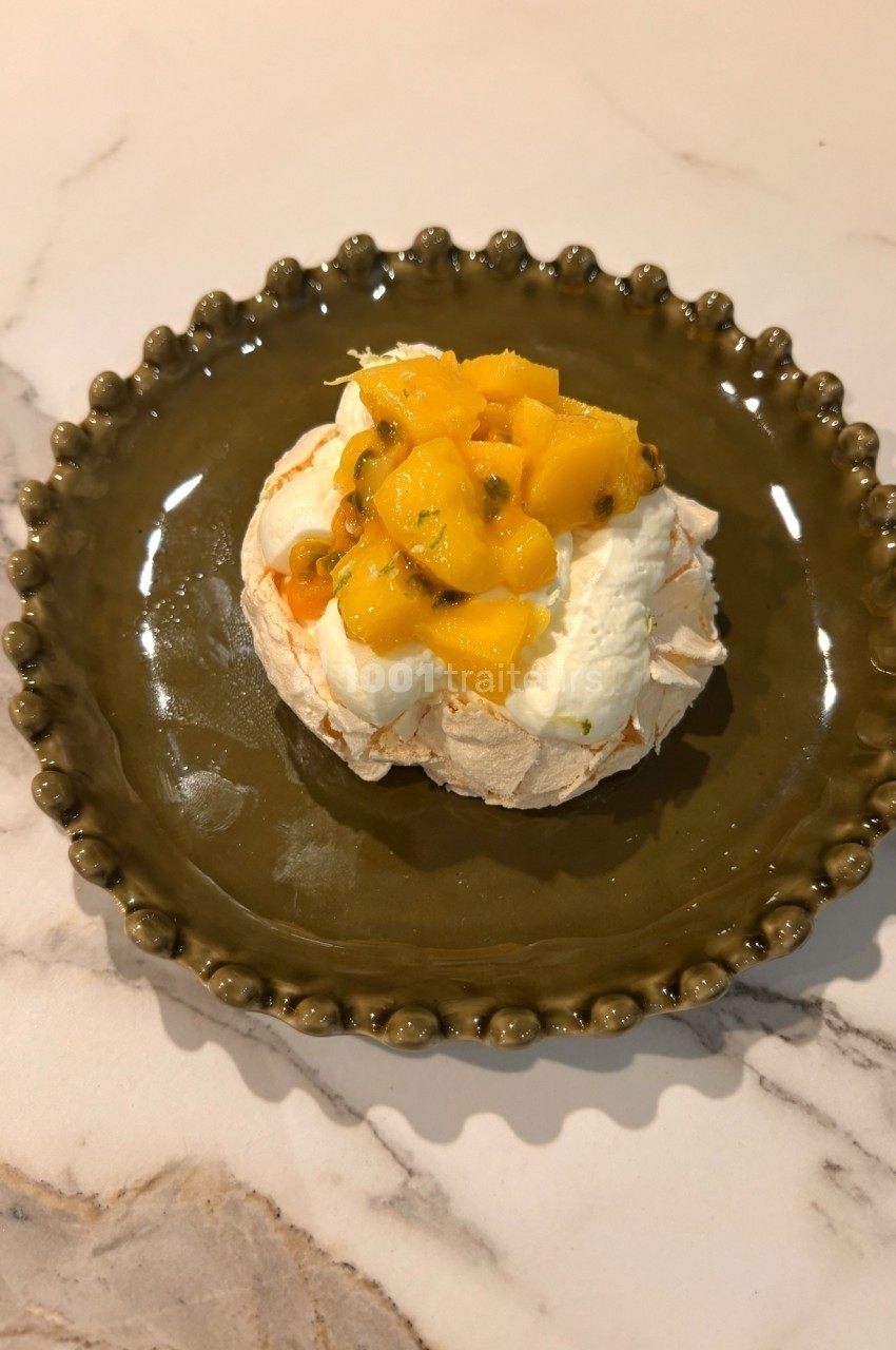 Dessert pavlova garni de crème fouettée, morceaux de mangue et fruits de la passion, présenté sur une assiette verte.