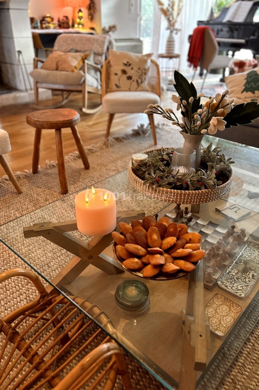 Table basse en verre décorée avec une bougie allumée, un plat de madeleines et un vase de fleurs, dans un salon chaleureux.
