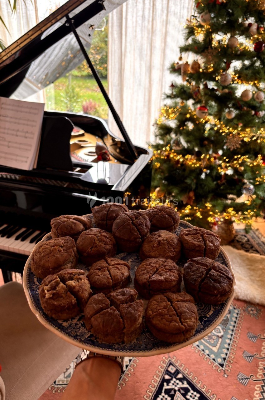 Assiette de muffins au chocolat tenue devant un piano à queue et un sapin de Noël décoré dans un salon lumineux.
