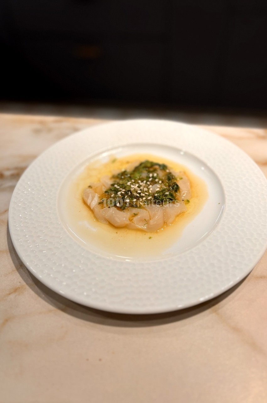 Assiette de carpaccio de poisson cru, nappé d'une sauce verte et parsemé de graines, sur une table en marbre.