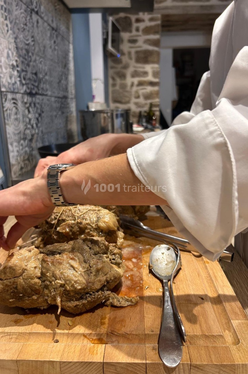 Un cuisinier attache une pièce de viande cuite sur une planche en bois, avec des ustensiles posés à côté.