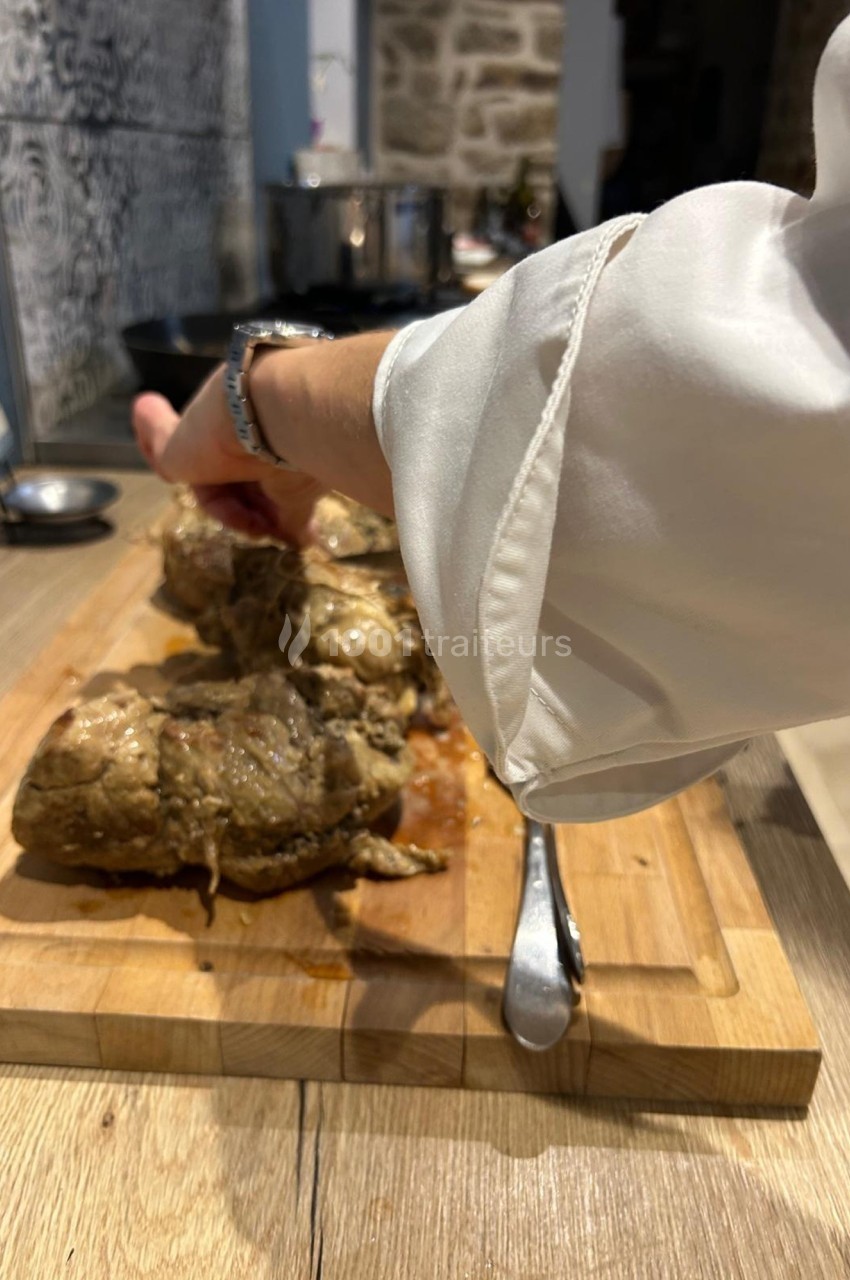 Une main en chemise blanche prépare des morceaux de viande cuits sur une planche en bois dans une cuisine.